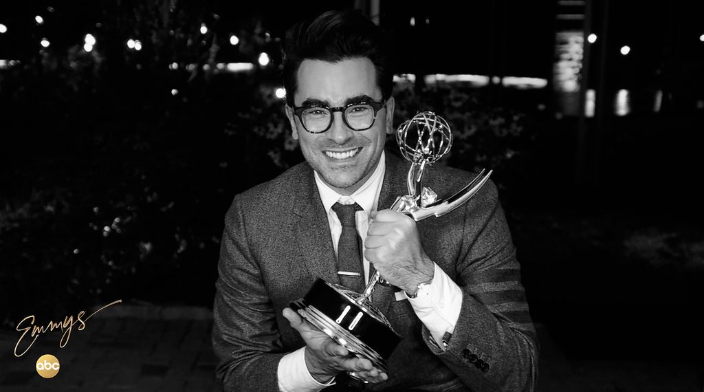 Black and white photo of Dan Levy holding his Emmy with two hands at the 72nd Emmys in September of 2020. Levy wears a suit and tie and smiles wide in his thick-framed glasses. Branded overlay reads “Emmys, ABC” in the bottom left corner.