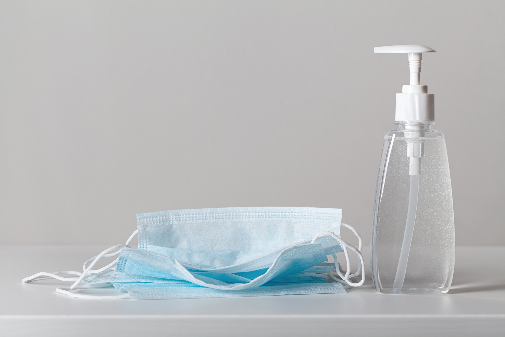 Photo of a pile of blue medical face masks on the left and a bottle of hand sanitizer on the right; they both rest on a white surface.