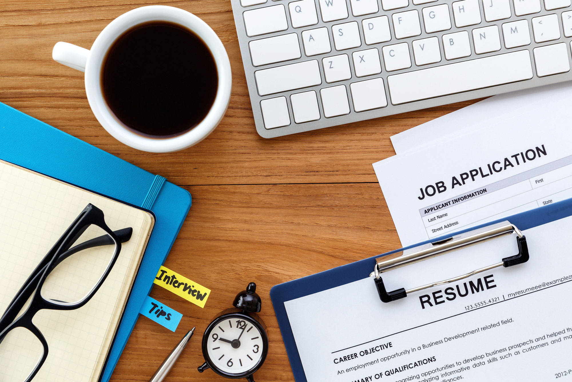 A photo of a desk cluttered with job applications, resumes, a keyboard, a pair of glasses, and a cup of coffee among other things.