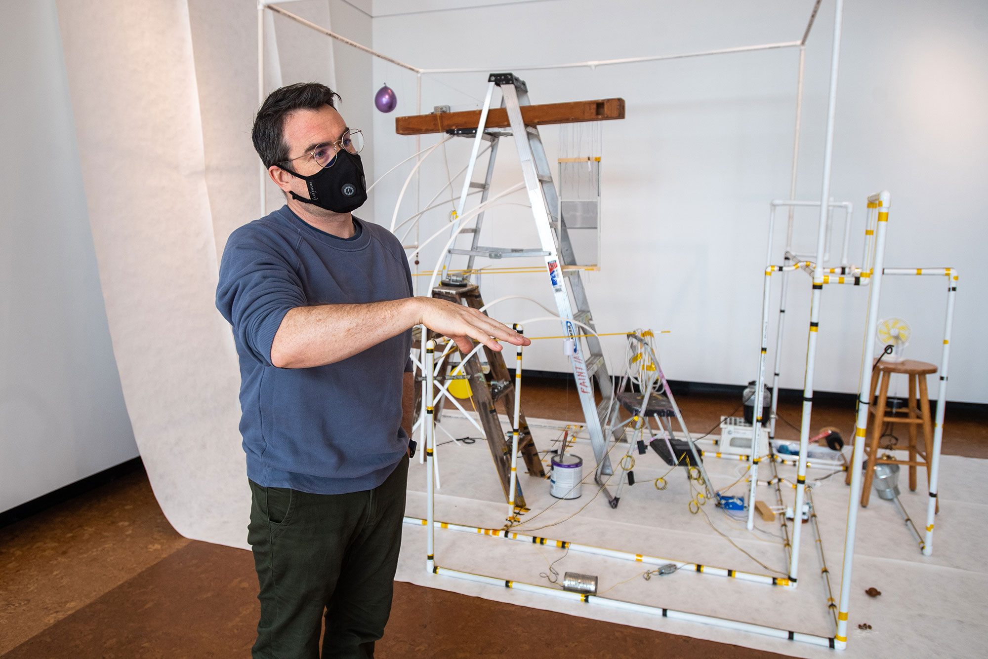 Photo of CFA’s David Snyder, in a blue long sleeve and black face mask, at the 2021 MFA Thesis Sculpture Exhibit. He stands in front of an untitled piece by Aris Hu (MFA’21), which consists of a structure made of pipes that contains different sizes of ladders and stools.