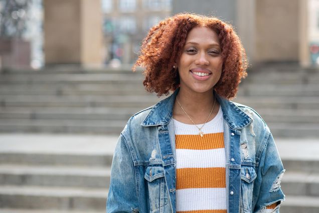Photo of Nyah Jordan (CGS’20, COM’22) standing in front of the steps in Marsh Plaza. She smiles, has curly shoulder-length reddish hair, and wears a jean packet and gold and white striped sweater.