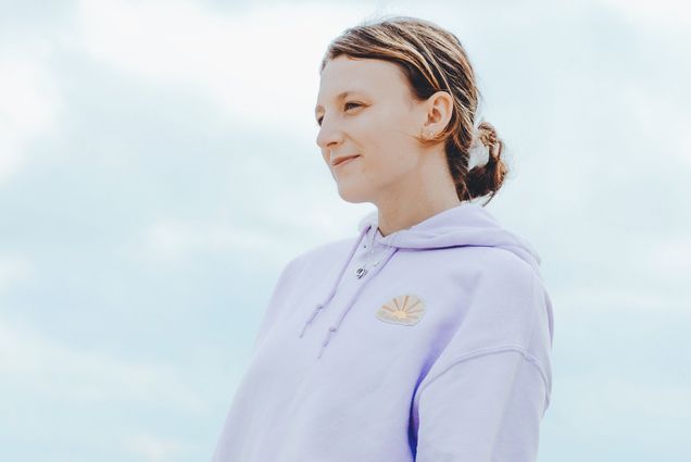 Photo of Kate Silvestri (Questrom’23) modeling the light purple Lotus hoodie, one of the pieces in her Radiate clothing line. Behind her, a gray sky and body of water are seen.