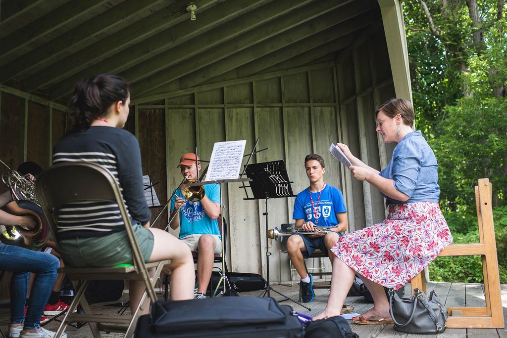A small group of young musicians practice in a structure that resembles a wooden shed and an outdoor stage; in the background, lush green trees are seen. At the front of the structure, an instructor with pages in her hand seems to conduct the students in front of her, who either play their horns or wait for their turn with their instruments in their laps.