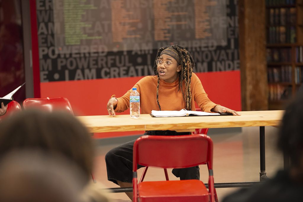 Photo of Ireon Roach (CFA’21) in The Jar’s reading of Alma by Benjamin Benne in 2019. She sits behind a table with a binder and water bottle in front of her and recites animatedly.