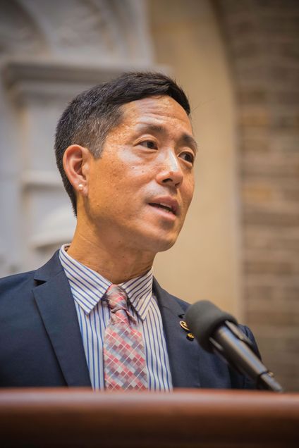 Photo of Kei Koizumi in a patterned tie and suit speaking into a microphone.