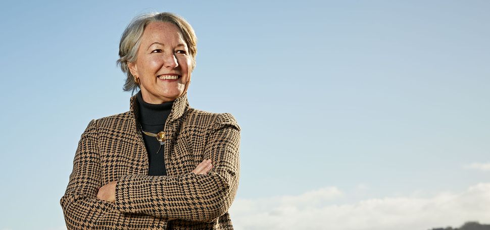 Photo of Susanne Daisley Lyons, smiling and standing in a striped blazer on top of a hill with her arms crossed. Behind her is a light blue sky and city skyline.