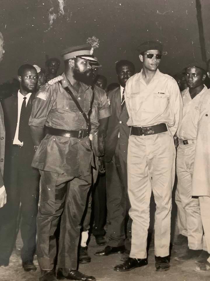 Chukwuemeka Odumegwu-Ojukwu (left), president of the short-lived state of Biafra, and Louis Austin Chude-Sokei (center, with sunglasses), a high-ranking Air Force major, and one of the leaders of the Biafran rebellion, stand in military dress at the front of a group of people.