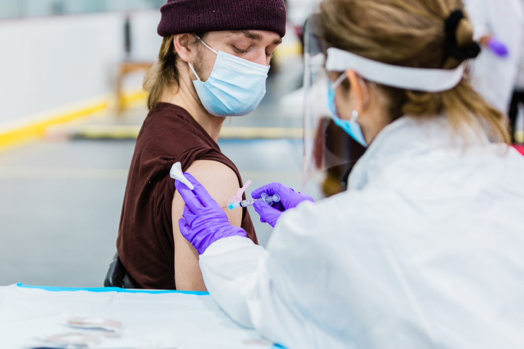Photo of Ian Forcey, in a brown beanie, looking down at his shoulder, as a medical provider in a face shield, mask and purple gloves gives him a vaccine.