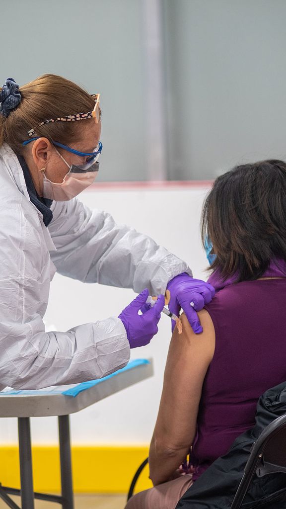 Photo of Terry Del Prato, RMA, in a white lab coat and purple glove, giving Yolanda Barros, seen from behind, with her jacket off one of her shoulders, receiving the Moderna COVID-19 vaccine January 20 at the vaccine clinic at FitRec.