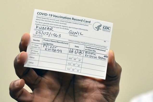 Photo of someone holding up their vaccination card. It reads “COVID-19 Vaccination Record Card….Last Name Kumar, First Name Sunil,” and other personal information. They received the Pfizer vaccine. The background is beige.