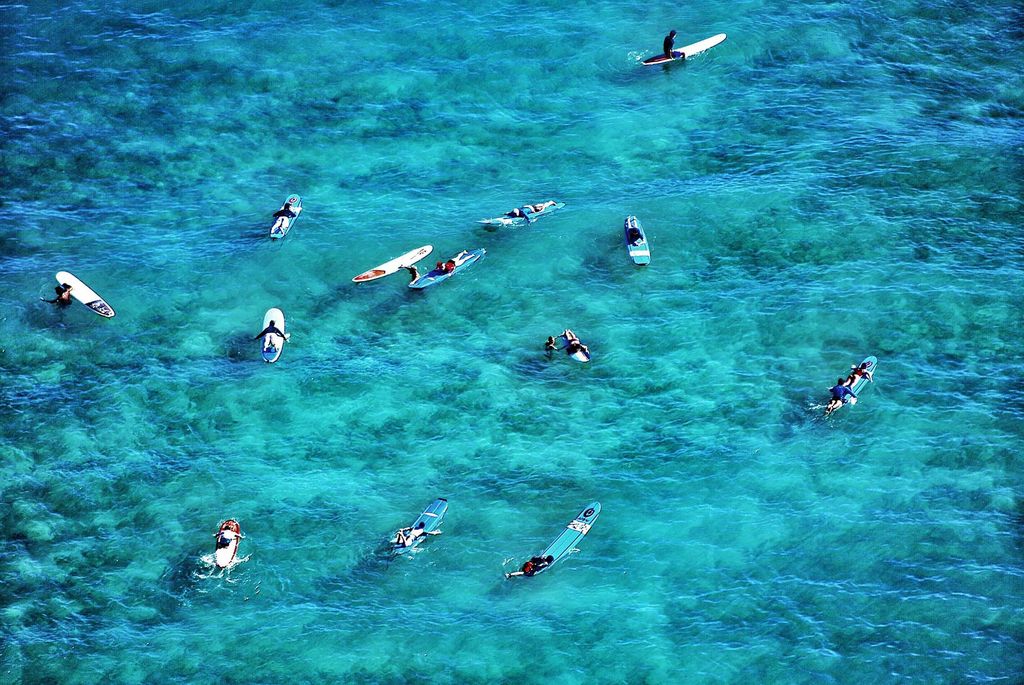 Photo taken in Waikiki, HI, an aerial shot of surfers catching waves in light blue, clear waters.