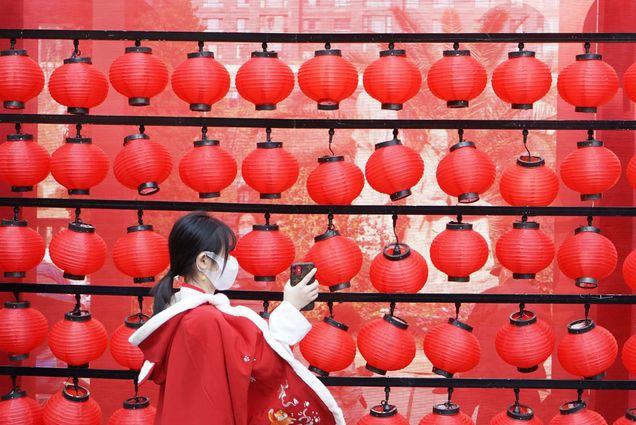 Photo called “New Year from Anywhere” in which a young girl holds up her phone towards a wall of red lanterns; she wears a red coat with an intricate design.