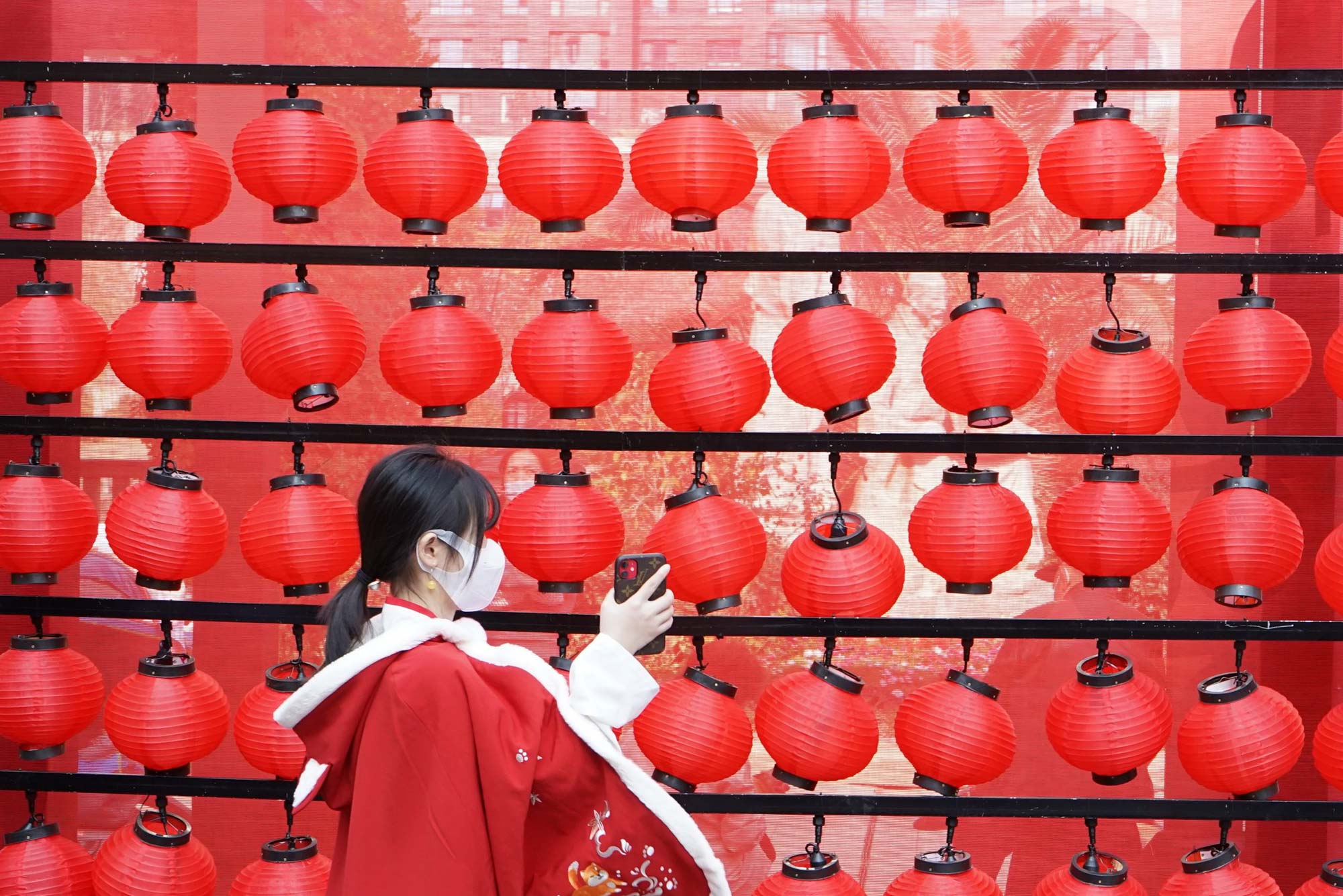 Photo called “New Year from Anywhere” in which a young girl holds up her phone towards a wall of red lanterns; she wears a red coat with an intricate design.