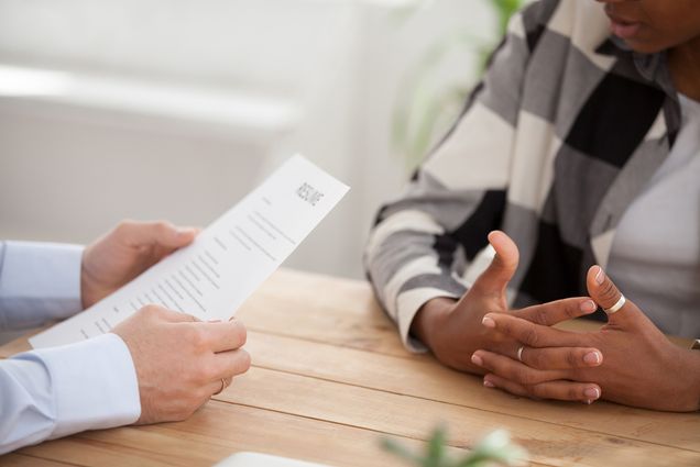 Photo of two pairs of hands resting on a table, on the left, a person with a resume in their hands interviews a job candidate on the right.