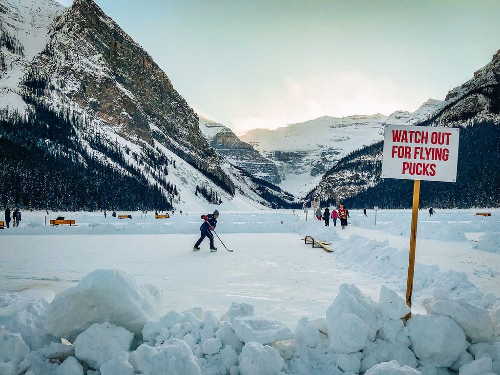 Photo of people playing ice hockey on a frozen lake where a section of the snow has been shoveled to expose the ice. A sign in the foreground reads "Watch out for flying pucks!" A snowy mountainous landscape is seen in the background.