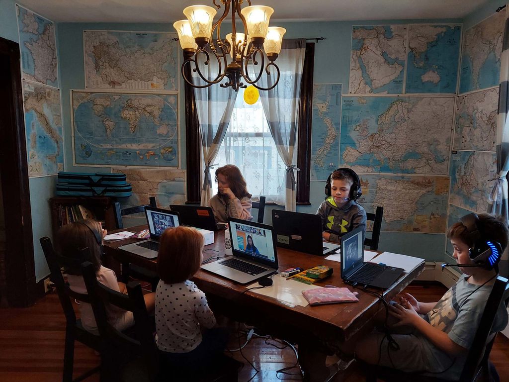 Photo called “Family from Anywhere” in which four young children sit at a kitchen table on zoom school with their mom. The room is plastered with maps floor to ceiling and a bright light fixture hangs from the ceiling.