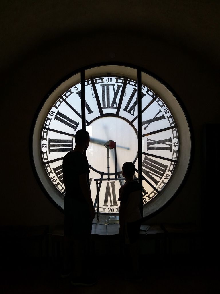 Photo of two people, one adult and one child-sized person, standing in front of a clock on the inside of the tower. The clock has large roman numerals and the people are seen in silhouette.
