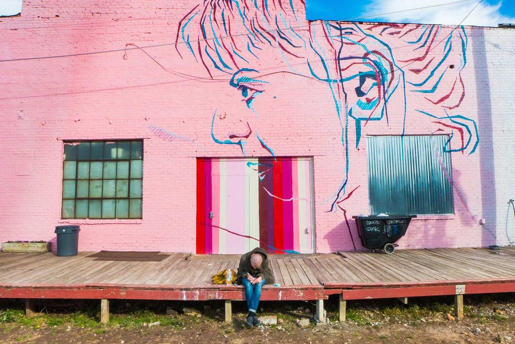 Photo of a person hunched over, sitting at the edge of a wooden platform, outside of a building painted bright pink with a mural of someone's face in profile.