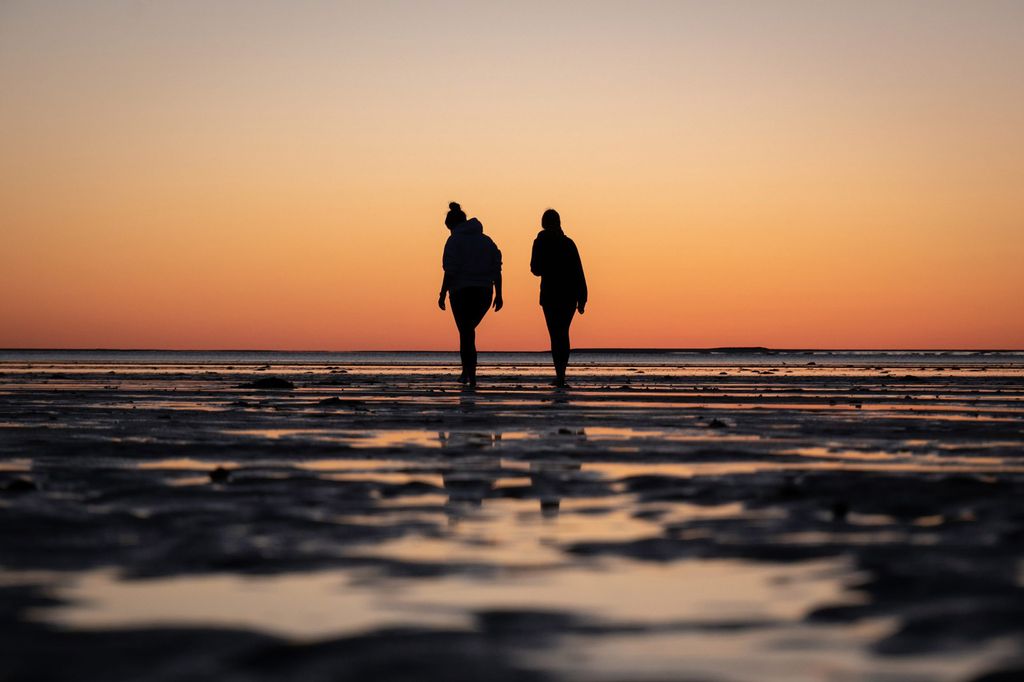 Photo of two people in silhouette walking down the shore of a beach at sun set, the sky is orange.