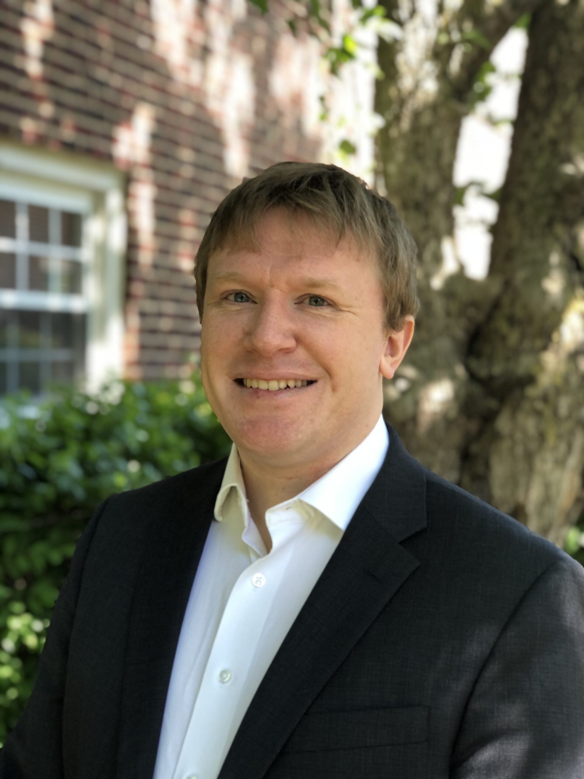 Portrait of Soren Hessler, smiling, in a black suit jacket and white shirt standing outside in front of a tree and brick building.