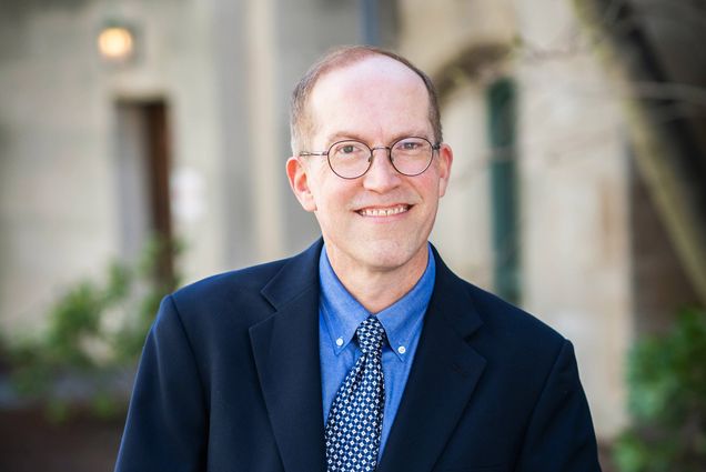 Headshot of Metcalf Award winner David Sullivan, a CAS master lecturer in computer science. He wears a blue suit and tie and has rounded glasses; he smiles as he leans on a half-wall outside.