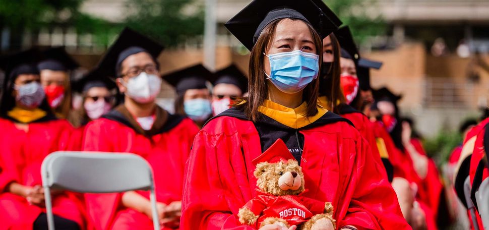 A graduating student wearing a black graduation cap and red gown holds a BU teddy bear while watching the 2021 BU Commencement for undergraduate students on Nickerson Field.