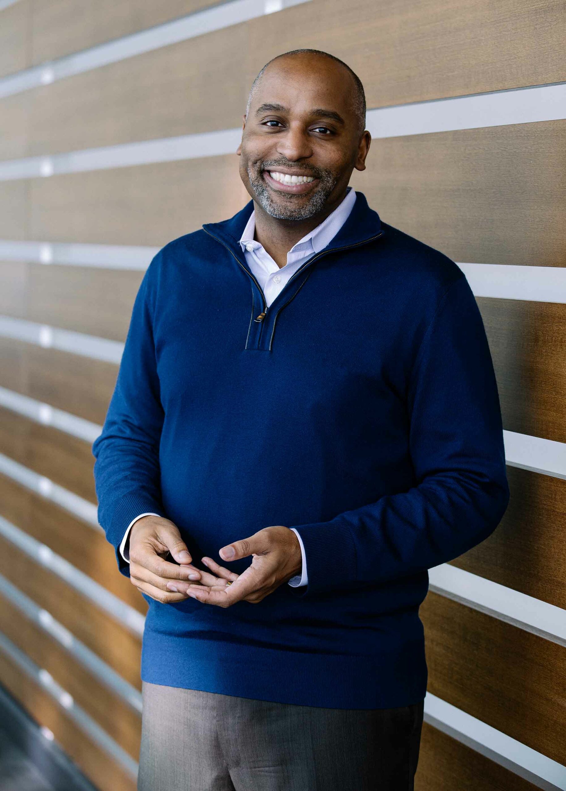 Portrait of Harvey Young, a middle-aged Black man. He wears a blue quarter zip sweater and smiles. He leans against a wall with beams running parallel to the ground.
