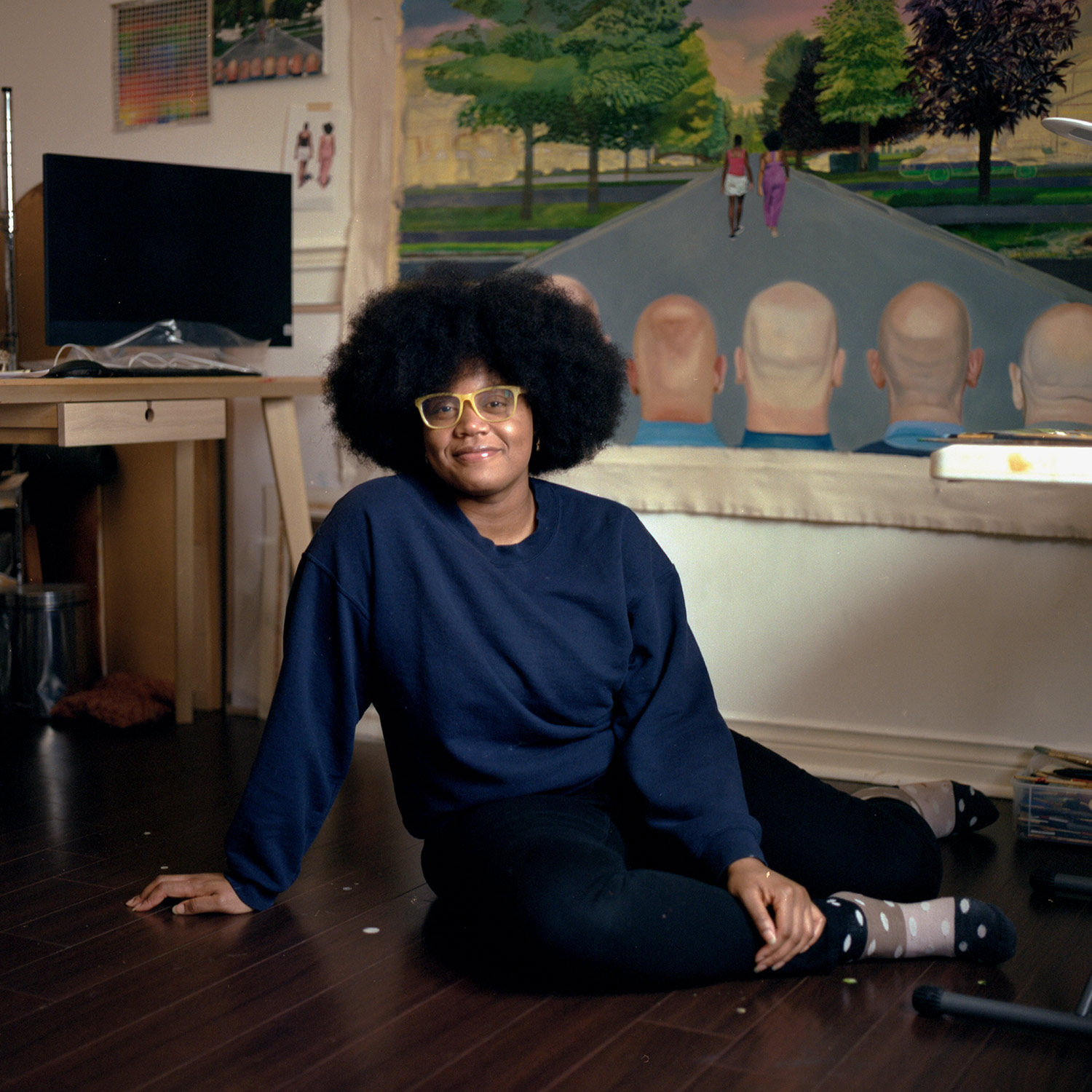 Photo of Shantel Miller (CFA’21) sitting on the ground and smiling. She wears a blue sweatshirt, black pants, polka-dotted socks, and yellow glasses frames. Behind her, a desk with a computer is seen and a painting that shows the back of the heads of a group of white men as two young black women walk down the street.
