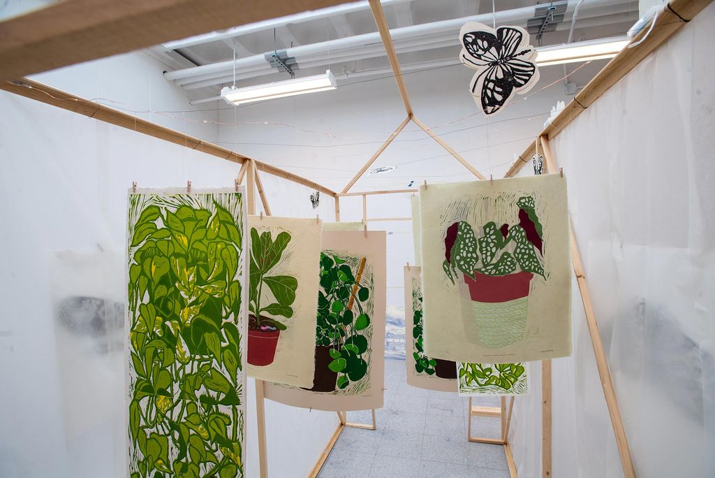 Photo of hand-built, wooden and plastic greenhouse made by Manon Michel (CFA’21) in which reductive linocut depictions of plants in green hang from the rafters.