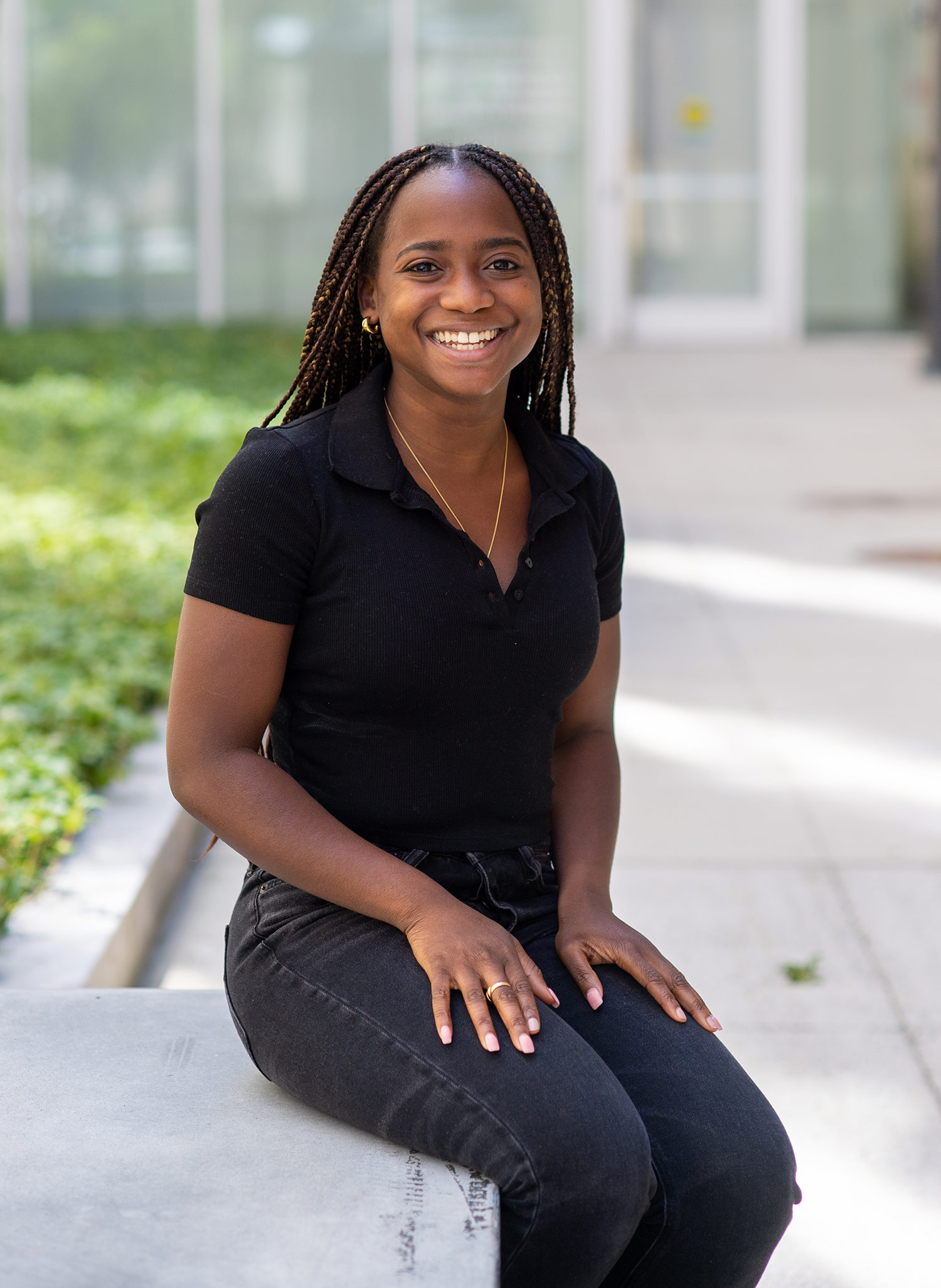 Photo of Archelle Thelemaque (COM’21) in black jeans and a black shirt sitting on a granite bench outside of one of buildings on campus. She has long brown braids and rests her hands on her lap as she smiles.