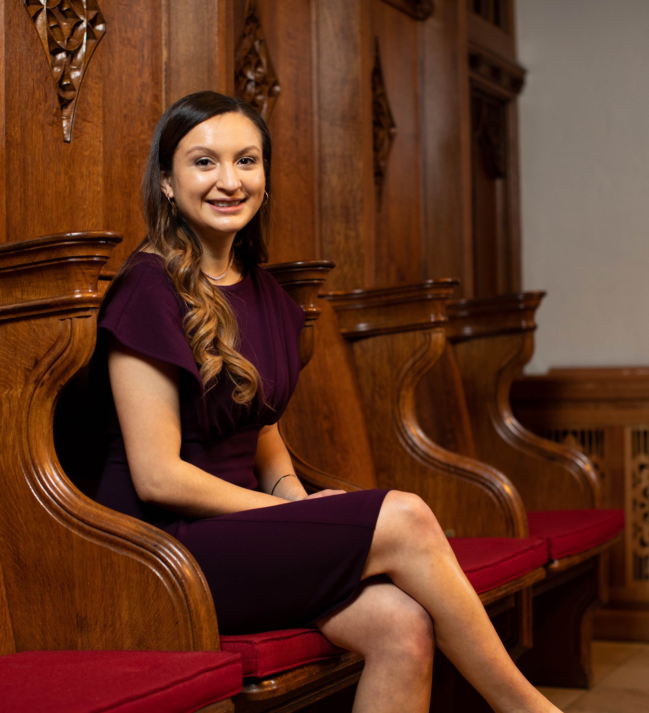Photo of Heidi Santa Cruz in a dark purple dress, sitting on an ornate wooden bench with red cushions, with her legs crossed. She smiles, and has long brown hair.