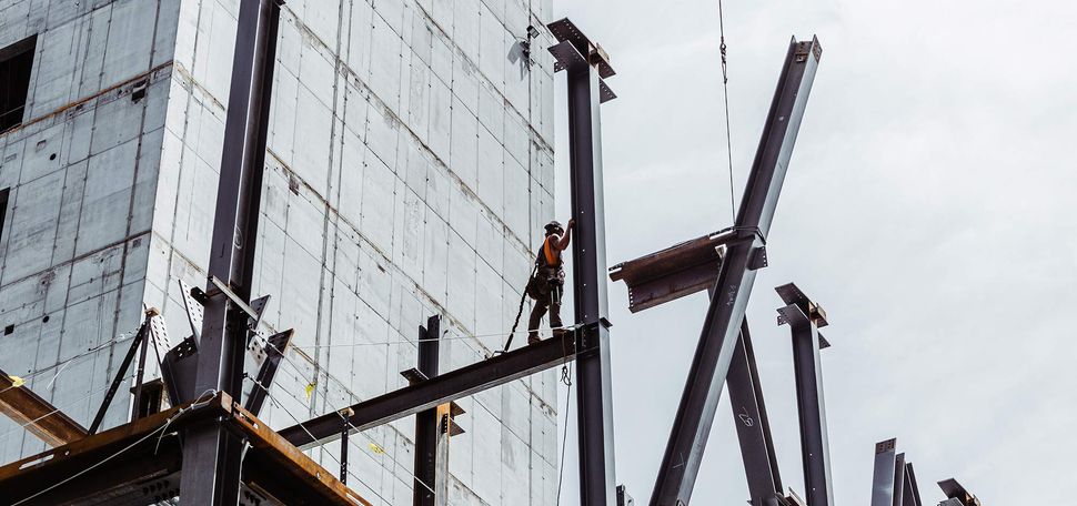 Photo of a construction worker standing on the large steel beam scaffolding of the future Center for Computing & Data Sciences building. The sky behind them is light blue.