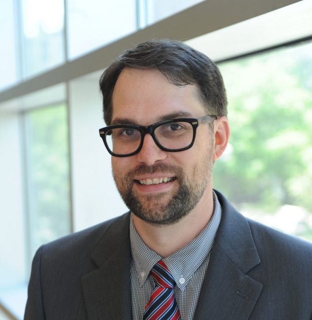 Headshot of Mark P. Newton, who wears glasses, a gray suit and a striped tie, standing in front of a window.