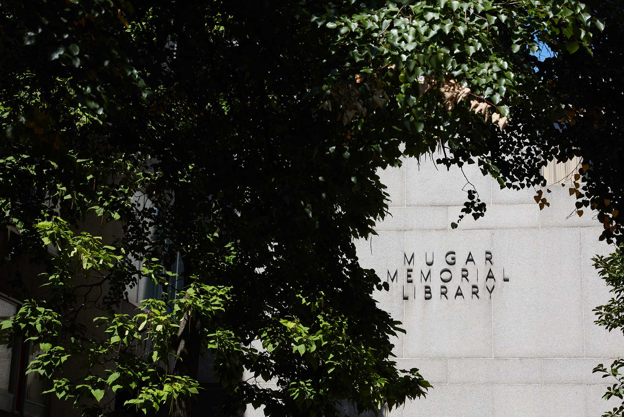 Photo of the entrance to the library as seen through a break in the trees. A sign that reads “Mugar Memorial Library” hangs on a gray, granite wall.