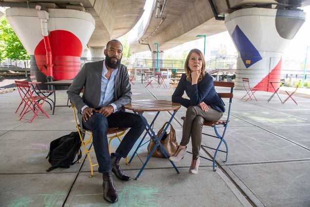 Daren Bascome, in gray jacket at left, and Chris Needham (CAS’86), wearing a blue blazer with her hand on her chin at right, sit at a colorful outdoor table under and overpass.