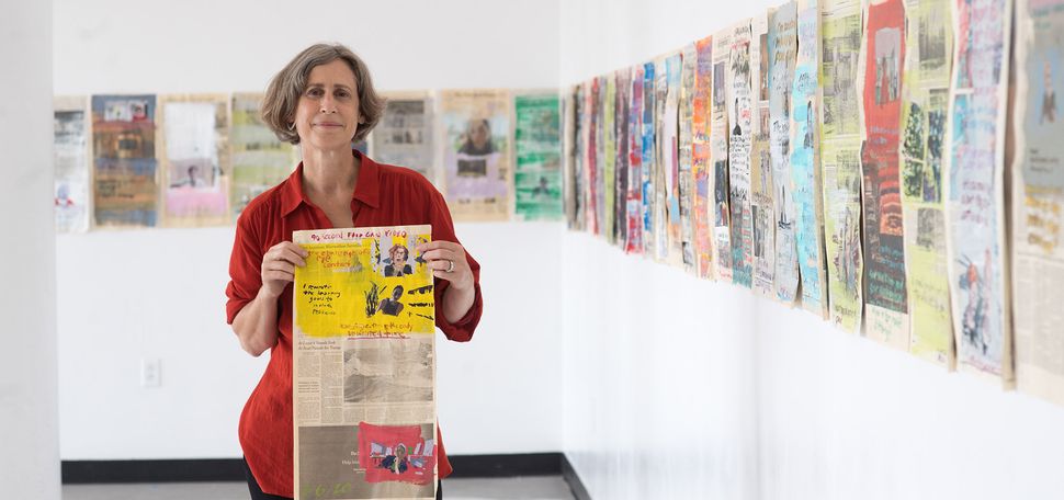 Photo of CFA SVA Director Dana Clancy in a red button down looking towards the camera as she holds a painted page of the NYTimes in a gallery of her work.