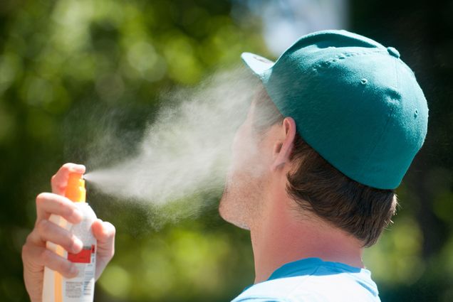 Photo of a young person in a green baseball cap spraying their face with sunscreen.