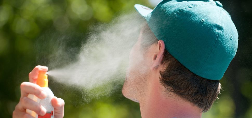 Photo of a young person in a green baseball cap spraying their face with sunscreen.