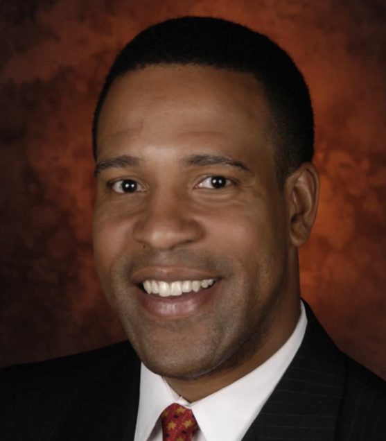 Headshot of K. Matthew Dames wearing a black suit and red tie, smiling, in front of a reddish background.