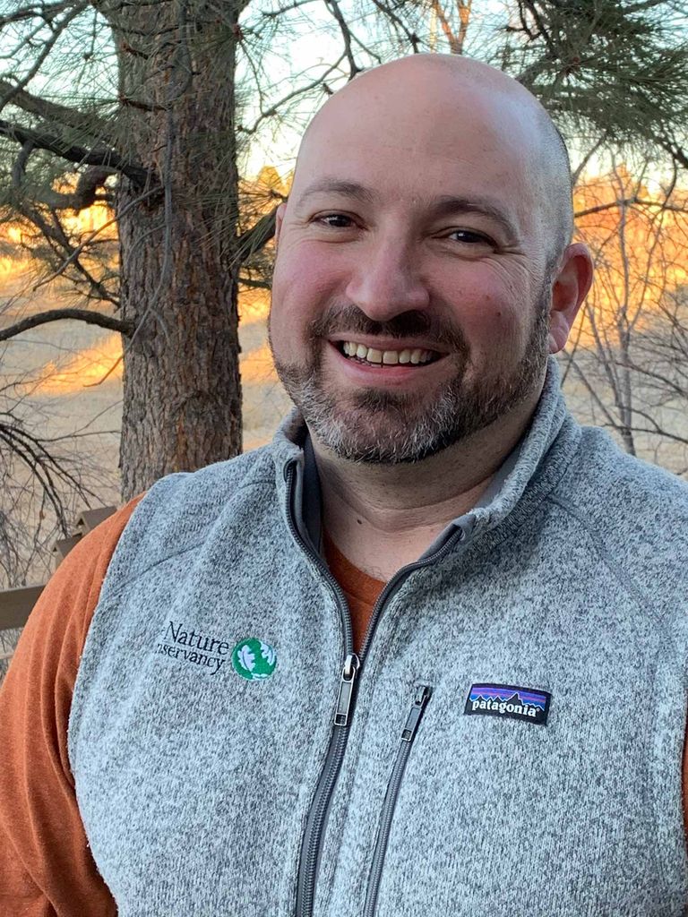 Photo of Jesse Gallun in a gray patagonia vest and mustard colored shirt, smiling, with trees and a mountain in the background.