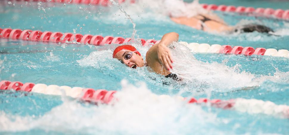 Photo of Julimar Avila (Sargent’19) during a swim meet at BU; in the photo she swims between lanes lined with red and white plastic lines and wears a red swim cap and googles.