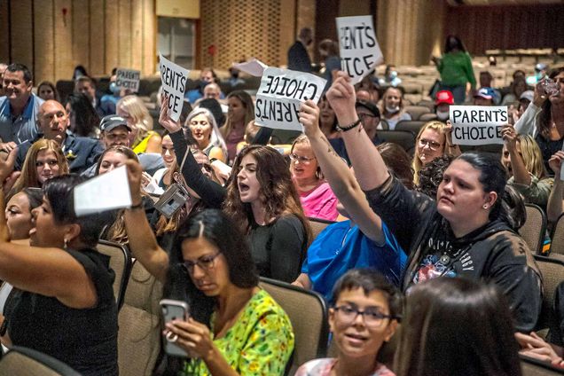 Photo of a parents holding signs that say “Parents Choice” at a Pennsylvania school board meeting on Aug. 25. They do not wear masks, and some appear to be yelling. Some children are also seated in the auditorium.