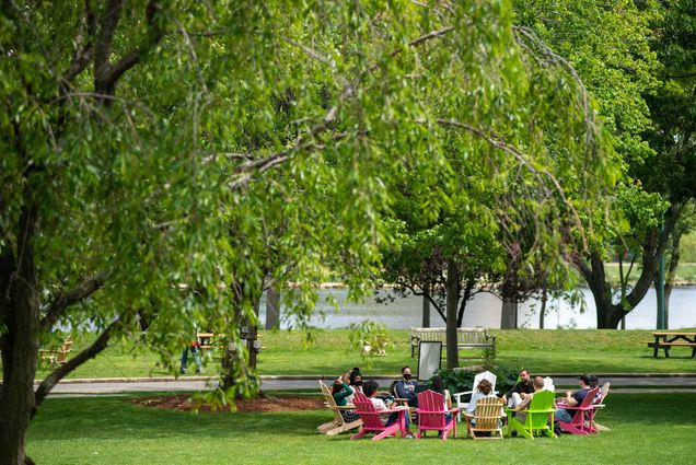 A class is lead in discussion sitting in a circle of colorful adirondack chairs beneath a tree on BU Beach, Boston University Charles River Campus.