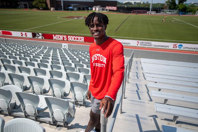 Photo of Tyler Johnson (COM’25) standing on a staircase in the seats along the men’s soccer field. He smiles, and wears a red Boston University under armor long-sleeve shirt, and leans against the railing. The bright green turf field is seen behind him.