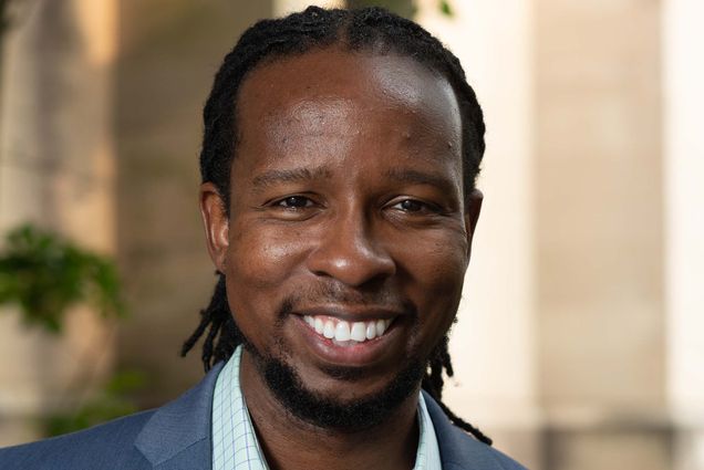 Photo of Ibram X. Kendi smiling. He has shoulder length locs that are pulled back, and he wears a blue suit, colorful pocket square, and light blue button up shirt. The portrait was taken outside, a building and a tree are blurred in the distance.