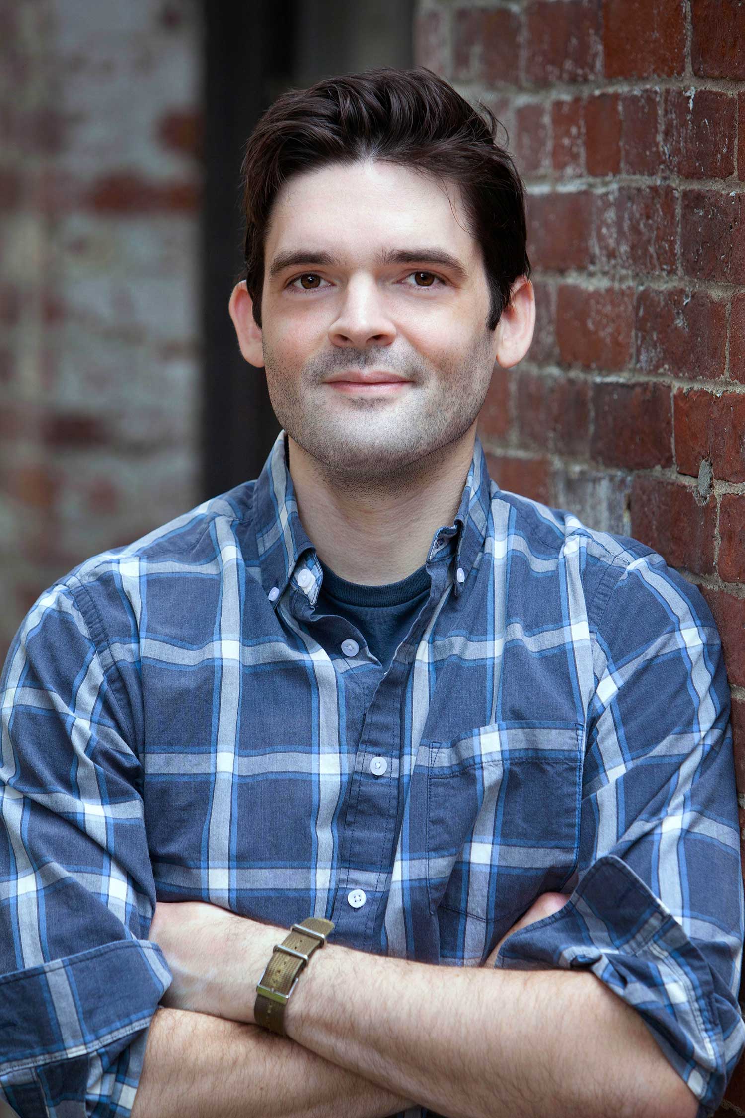 Headshot of Paul Coleman (COM’21) wearing a blue and white plaid shirt with his arms across his chest. He smiles slightly and leans against a brick wall. He has dark brown hair.