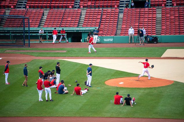 Pitcher Chris Sale practices on the mound while team mates watch. Red sox team members dressed in red and blue stand behind the mound, and Sale is seen mid pitch. The photo was taken during a practice on June 11, 2021 and the red seats in the stands are empty.