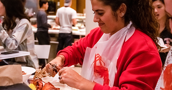 Photo of Emma Moneuse (CGS ’22), in a red sweatshirt and white, plastic lobster bib, digging into her lobster at Marciano Commons Dining Hall. Other students are seen in the background.