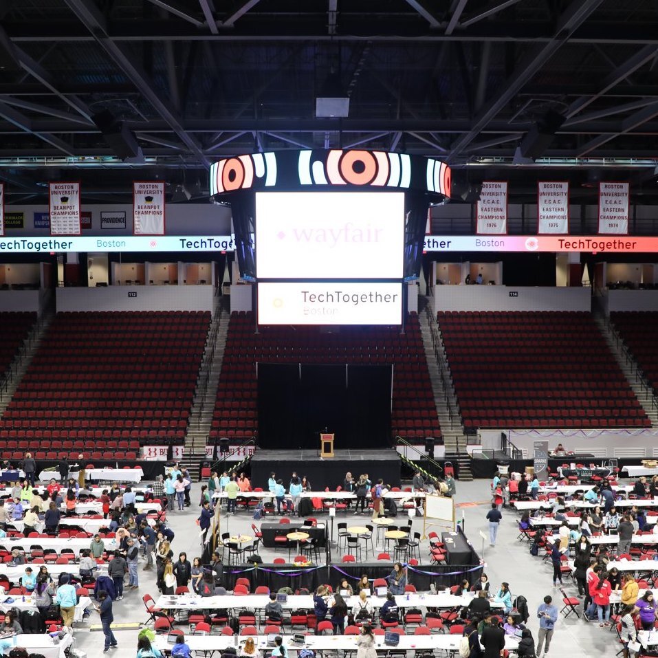 Photo inside what appears to be Agganis Arena during TechTogether Boston 2019-2020, one of the last in-person hackathons TechTogether helped organize before the coronavirus pandemic. People walk around tables with red chairs on the floor.