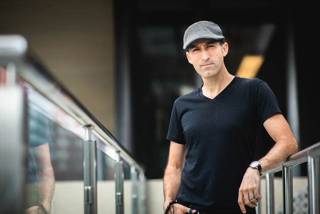 Photo of David Delmar Senties, wearing a gray hat, and a black t-shirt, as he leans across a clear balcony with a silver railing.